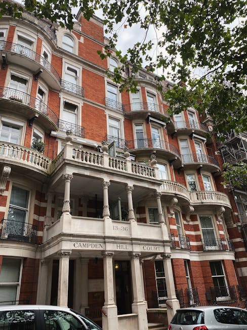 A multi-storey residential building with red brick façade, white decorative stonework, and curved bay windows visible on each floor. The building features ornate balconies with black wrought iron railings and a prominent entrance supported by classical columns, inscribed with the address '13-36 CAMPDEN HILL COURT'. The scene is shaded by large leafy trees in the foreground, with a clear sky overhead. In front of the building, a paved area is visible with a few parked cars. This image depicts the exterior of a typical London property, relevant to house removals and home relocation services offered by West Kensington Man and Van, supporting efficient furniture transport and packing and moving activities in the W14 postcode area.