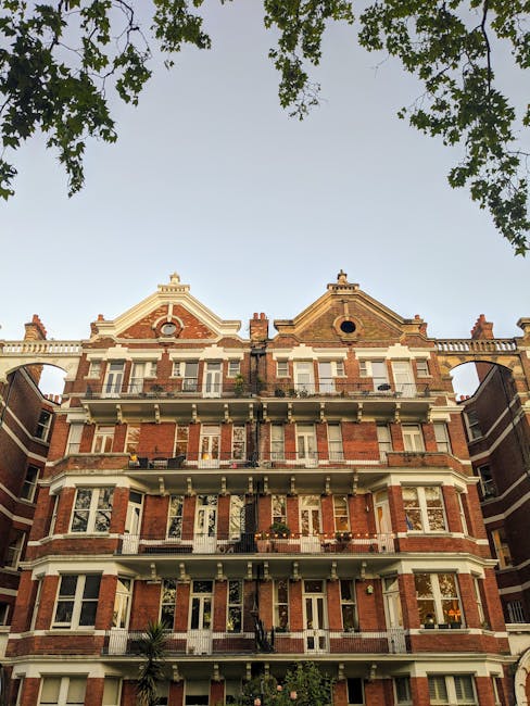 The image depicts a multi-storey Victorian-style brick building opposite a street, with a clear sky overhead. The building features ornate architectural details, including decorative white trim, bay windows, and small balconies with black railings on several floors. There are large, rectangular window openings, some with partially open blinds, and a circular attic window near the roof peak. The perspective is from ground level looking up, with tree branches and leaves partially framing the top of the image. The scene suggests an urban residential area in West Kensington, where house removals and furniture transport services by West Kensington Man and Van could facilitate home relocation projects, involving the careful packing and loading of items through the building’s entrance or rear access.