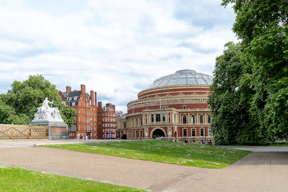 A wide view of a large, historic circular building with a domed roof made of metal and brick exterior, situated in an urban park environment. In the foreground, a paved pathway runs through a grassy area with patches of white flowers. To the left, a white stone sculpture of a seated figure with an arm raised is mounted on a pedestal, partially obscured by lush green trees that extend along the right side of the image. Behind the sculpture, apartment buildings constructed from red brick with multiple chimneys and windows line the background. The scene is lit by natural daylight, with a partly cloudy sky overhead. The image captures the peaceful setting ideal for home relocation or furniture transport services, as provided by West Kensington Man and Van, with an emphasis on moving logistics in West Kensington.