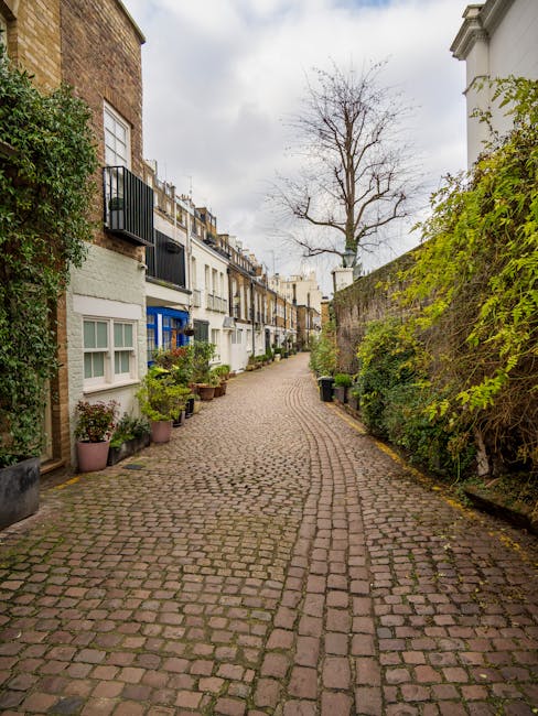 A narrow cobblestone street in West Kensington lined with terraced houses featuring ground-floor windows, some with flower pots and potted plants outside. On the left, there are white and blue painted facades, with balconies and small drainpipes, while the right side has a high brick wall covered partially with green foliage. In the background, a leafless tree stands against a cloudy sky, indicating late autumn or winter. The street appears quiet and clean, with no visible moving vehicles or furniture. This setting provides a typical example of an urban residential area suited for house removals and home relocation services, where professional movers like West Kensington Man and Van might carry out furniture transport or packing and moving activities in accordance with local disposal and skip rules.