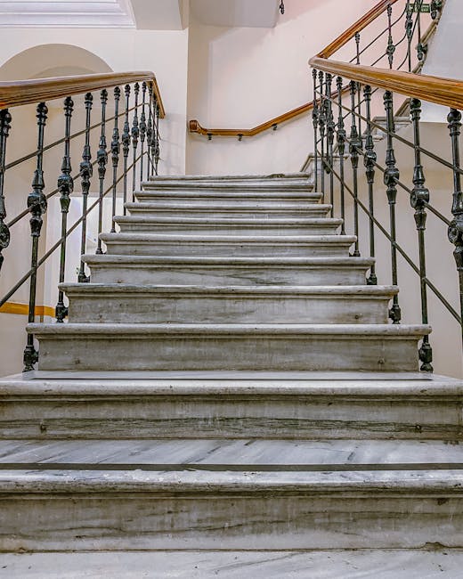 A marble staircase inside a residential building with multiple steps leading upward, featuring black metal railings with decorative balusters on both sides and wooden handrails mounted on the wall. Th