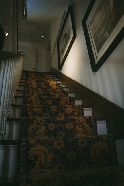 A marble staircase inside a residential building with multiple steps leading upward, featuring black metal railings with decorative balusters on both sides and wooden handrails mounted on the wall. Th