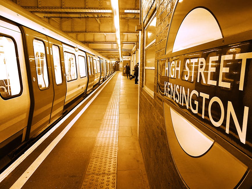 Inside the underground station at High Street Kensington, a modern, yellow-painted London Underground train is stationary alongside the platform. The platform surface features tactile paving strips ne