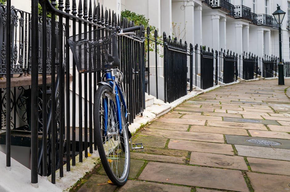 A blue bicycle with a black front basket is leaned against a black wrought iron fence along the sidewalk outside a row of white terraced houses with black railings and stairs. The bicycle’s kickstand is down, supporting its upright position, and the pavement is made of large, rectangular stone slabs with some moss or grass growing between them. The scene is outdoors during daylight, with natural lighting illuminating the area. Behind the fence, parts of the house entrances, including white steps and black iron railings, are visible. This setting depicts a residential street in West Kensington, where house removals and furniture transport often involve careful logistics, as seen in the image with the roadside fencing and outdoor environment. West Kensington Man and Van may sometimes facilitate such home relocation activities, requiring efficient loading and unloading processes.