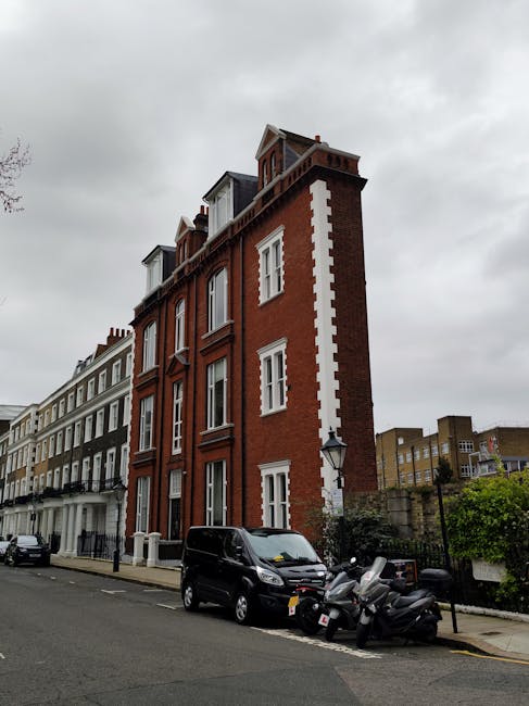 A multi-storey red brick building with white architectural details and large windows, situated on a residential street under an overcast sky. In front of the building, there is a black van and several motorcycles parked along the pavement, with some parked close to the building's entrance. The street appears quiet, with other similar residential buildings visible along the road. The scene relates to house removals and relocation services provided by West Kensington Man and Van, highlighting the urban environment where furniture transport and home moving activities commonly take place. The image captures a typical setting for loading and unloading during a house removal, with the building's exterior and parked vehicles emphasizing the logistical aspects of packing, moving, and transporting belongings in a city environment.