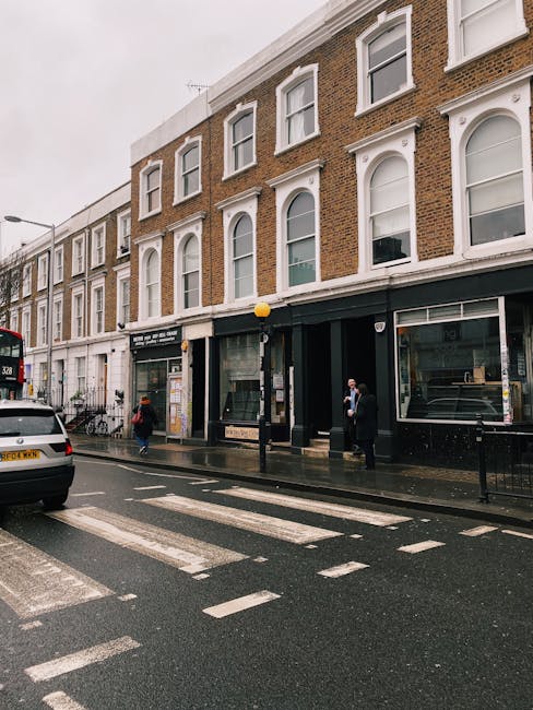 The image shows a street scene on Gloucester Road in West Kensington, featuring a three-storey brick building with large ground-floor windows and white decorative window frames on the upper floors. The pavement in front of the building is wet, indicating recent or ongoing rain, with some pedestrians walking across the zebra crossing. A black van from West Kensington Man and Van is parked on the street, partly visible on the left side, and a red double-decker bus is also seen further down the road. The ground-floor storefronts include glass doors and display windows, some of which are part of property units likely involved in residential or commercial use. A street lamp with a yellow spherical light is positioned near the building, and a few individuals are standing outside, possibly engaged in loading or unloading activities. The scene captures a typical urban moving environment where furniture transport, packing, and home relocation logistics are taking place, with the context suggesting a professional removals service in an active city neighbourhood.