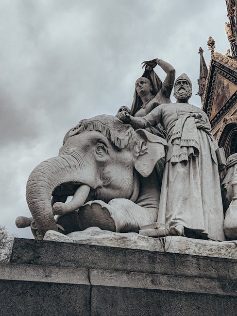 A large stone statue featuring an elephant with detailed carvings of wrinkles and textured skin, situated outdoors against a cloudy sky. On top of the elephant, there are three stone figures depicting historical or mythological characters, with the central figure seated on the elephant's back, holding an object, and flanked by two standing figures dressed in traditional attire with draped fabric and ornamental details. The sculpture is mounted on a rectangular stone base, which is part of a larger architectural structure with decorative elements visible at the upper right corner of the image. The scene is captured in natural daylight, highlighting the intricate craftsmanship and the monumental scale of the monument, which is located near a historic building or monument, as suggested by the ornate architectural features in the background. This type of sculpture is typical of commemorative or cultural monuments showing craftsmanship relevant to historical or cultural themes.