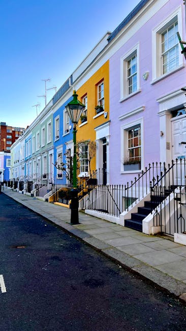 A row of colorful terraced houses in West Kensington, London, with pastel facades painted in shades of blue, yellow, pink, and purple. Each house features white-framed sash windows, small front gardens with black wrought iron railings, and steps leading up to front doors. A traditional black street lamp stands on the sidewalk in front of the row. The scene is illuminated by natural daylight, with a clear blue sky overhead. This exterior setting illustrates a typical residential street often associated with house relocation and moving services such as those provided by West Kensington Man and Van, which specialises in home removals near Olympia London. The image captures the charm of urban house transport logistics and the typical visual context for furniture transport and packing and moving activities within this neighbourhood.