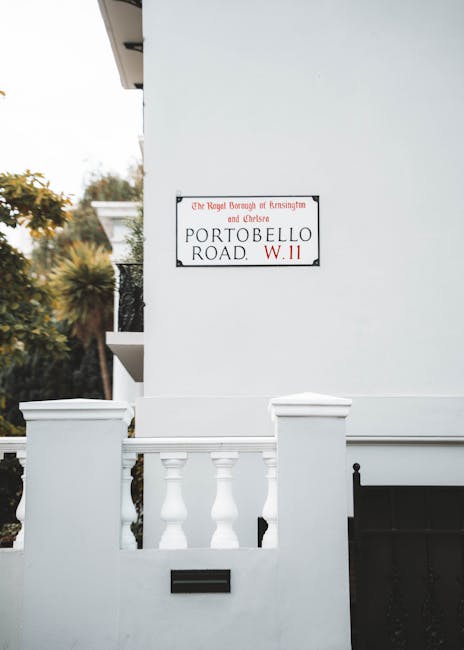 A white residential building with a small front porch featuring a white balustrade and decorative pillars, situated on Portobello Road in the Royal Borough of Kensington and Chelsea. The exterior wall displays a street sign indicating the location as part of the West Kensington area, with the text 'The Royal Borough of Kensington and Chelsea, Portobello Road, W.11'. The image captures the clean, modern façade of the property with a black mailbox on the wall below the street sign and an adjacent black gate partially visible to the right. The setting appears to be daytime with natural lighting, and in the background, blurred greenery and trees are visible, suggesting a quiet residential street. This scene relates to house removal and relocation services, highlighting the property’s exterior as part of the moving or packing process undertaken by West Kensington Man and Van, focusing on accurate visual representation of the home environment and surrounding context for furniture transport and home relocation activities.