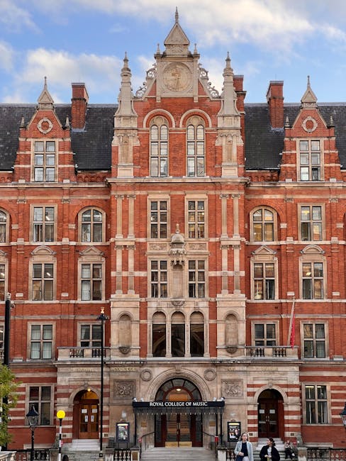 A large, historic red-brick building with intricate architectural detailing, including pointed gables, decorative stonework, and tall, narrow windows. The facade features a central clock and ornate spires at the roofline. In front of the building, there are two black lamp posts and a paved street with some pedestrians. The entrance to the Royal College of Music is visible at ground level, indicating it is located in an urban environment. The scene is well-lit with natural daylight, emphasizing the building's detailed exterior, which relates to house removals and moving services due to its prominent, structured appearance and urban setting. This image aligns with the context of professional relocation and furniture transport, as seen in home or institutional moves by West Kensington Man and Van.