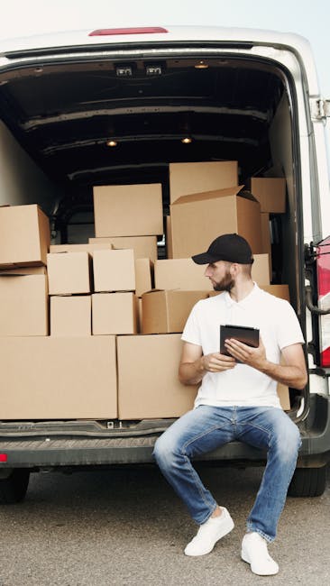A young man dressed in casual clothing, including a white t-shirt, jeans, and a black cap, is seated on the pavement next to the open rear of a van filled with cardboard boxes of various sizes, some sealed with packing tape and others open. Inside the van, the boxes are neatly stacked on the vehicle's floor and on custom shelving units, ready for a house removal or home relocation. Behind the man, the inside of the van is visible with a black ceiling and interior walls. The setting appears to be outdoors during daylight, with natural light illuminating the scene. The boxes are being prepared for transport, highlighting the packing and loading process typical of professional moving services. This image is associated with West Kensington Man and Van, a company providing removals and furniture transport services, as reflected in the context of the page about same-day man and van services in West Kensington.