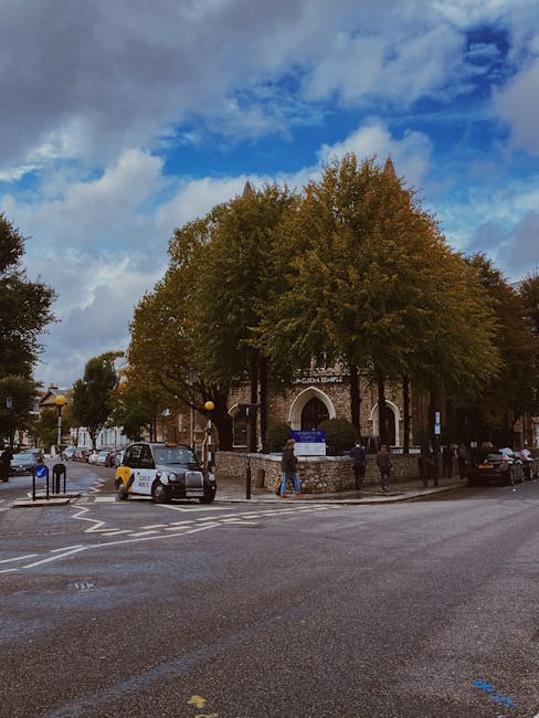 A daytime street scene showing the exterior of a brick building with an arched entrance, partially obscured by large trees with green and autumnal foliage. Several pedestrians are walking on the pavement and around the entrance, while parked cars and a small van are along the street. The sky above is partly cloudy with patches of blue visible. The area appears to be an urban environment consistent with park and building surroundings, and the scene suggests the context of home or office relocation activities. West Kensington Man and Van provides removals services, supporting efficient furniture and belongings transport within this setting.