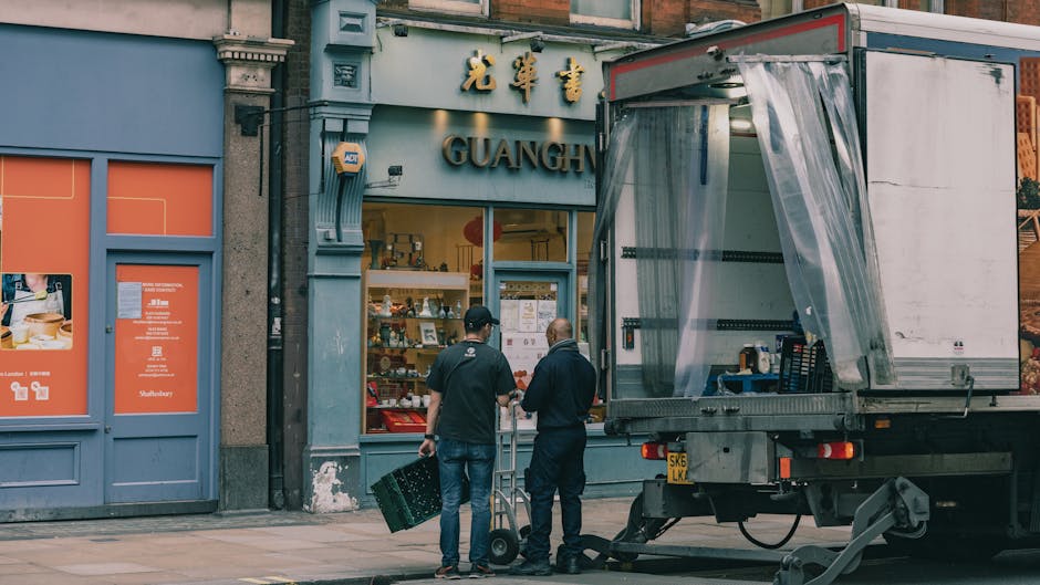 Two movers from West Kensington Man and Van, dressed in dark clothing, are positioned outside a shop on Kensington High Street, involved in the process of home relocation or furniture transport. One mover is holding a large black plastic crate, while the other is standing nearby with a handcart, both facing the shop. The shop's signage features Chinese characters and the word 'GUANGH', and the interior displays various items, likely souvenirs or gifts, visible through the large glass window. A moving truck with an open side door and plastic coverings on its loading ramp is parked directly outside, ready for loading or unloading goods. The scene is set on a pavement in an urban environment, with detailed elements including a small step at the storefront entrance, adjacent blue storefronts with orange informational posters, and the surrounding building facades. The lighting appears natural, suggesting daytime, and the overall context reflects professional packing, loading, and transport activities associated with house or office removals conducted by [COMPANY_NAME] as part of a comprehensive moving service on Kensington High Street.